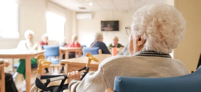 Elderly individuals seated at tables in a bright, communal room, engaged in conversation.