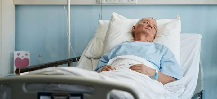 Elderly man in blue hospital gown lying in bed, looking upwards in a clinical room.