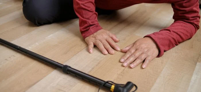 Close-up of elderly person’s hands resting on wooden floor next to a black walking cane.