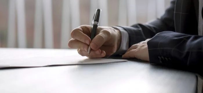 Close-up of a person in a dark suit signing a document with a black pen on a white table.