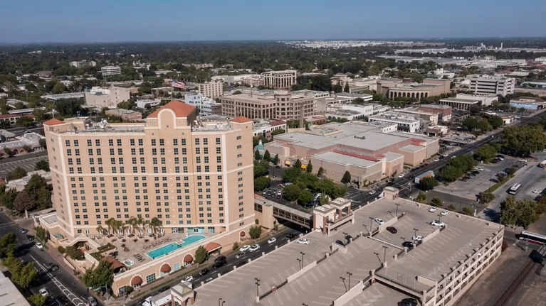 Aerial view of a large beige multi-story hotel with a swimming pool, adjacent parking structures, and surrounding urban buildings.