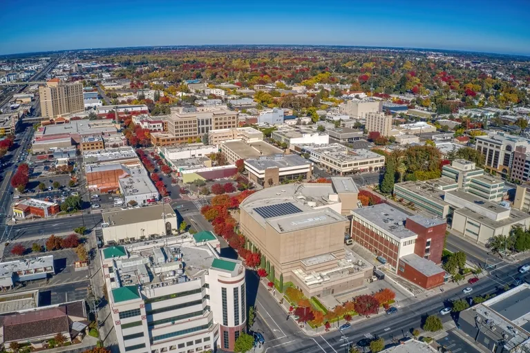 Aerial view of a mid-sized city with commercial buildings and autumn-colored trees under a clear blue sky.