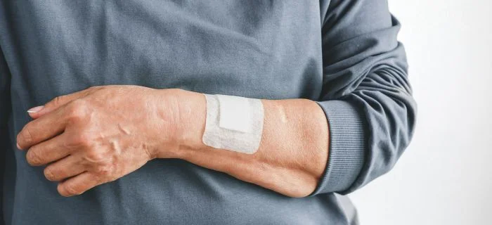 Close-up of an adult's forearm with a white adhesive bandage, wearing a gray long-sleeve shirt.