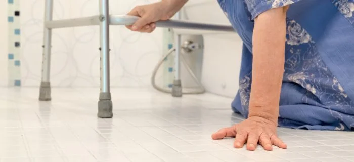 Elderly person in blue clothing gripping walker and supporting themselves on tiled floor.