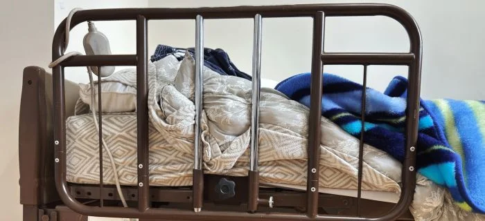Hospital bed with metal rails, white patterned bedding, and blue striped blanket.