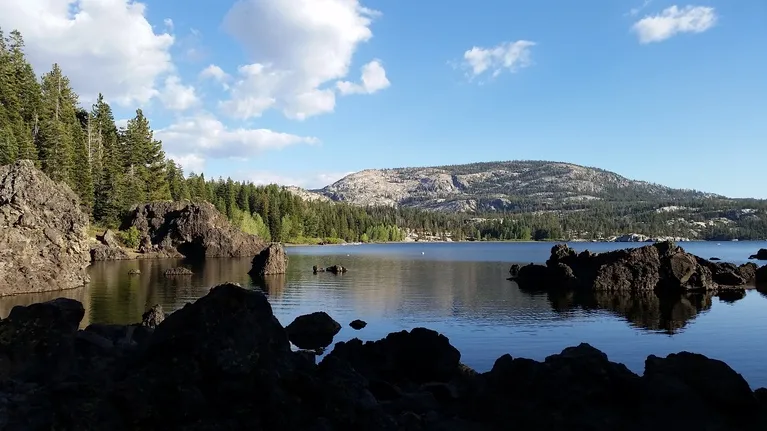 Calm lake surrounded by rocky formations, dense pine forest, and distant mountainous terrain under a partly cloudy sky.