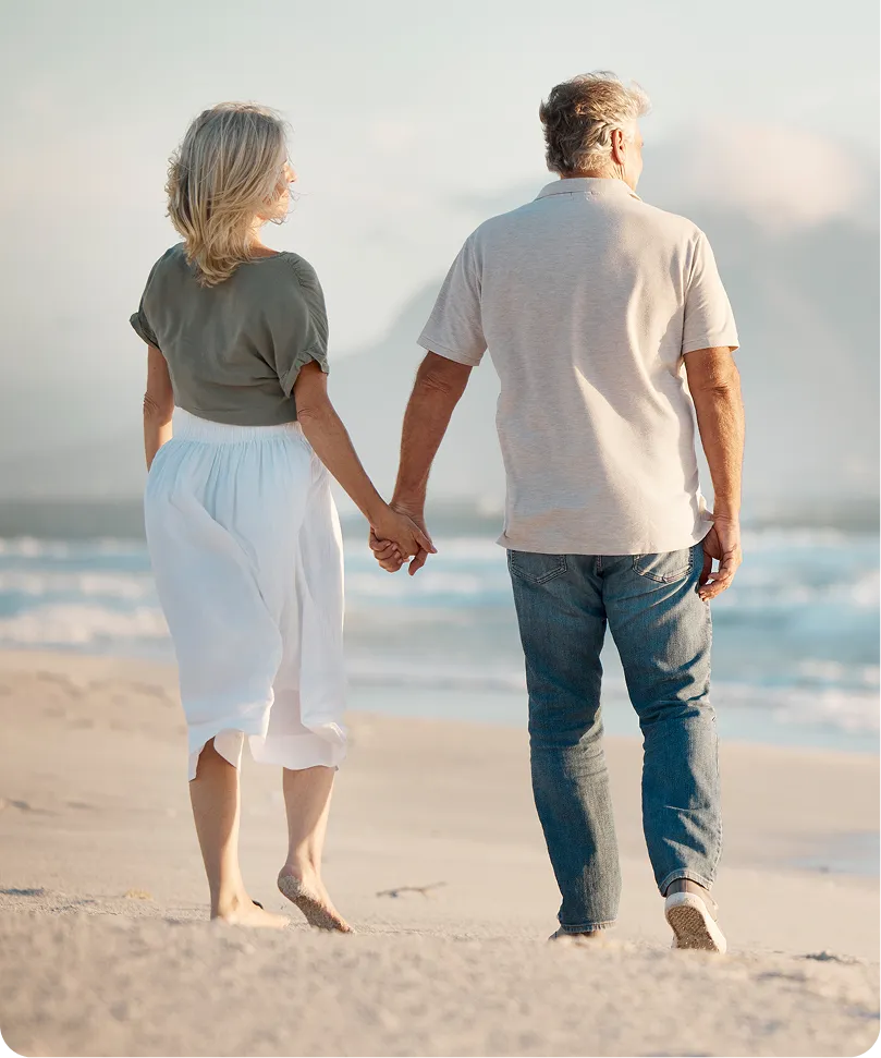 Mature couple holding hands, walking barefoot on a sandy beach near the ocean.