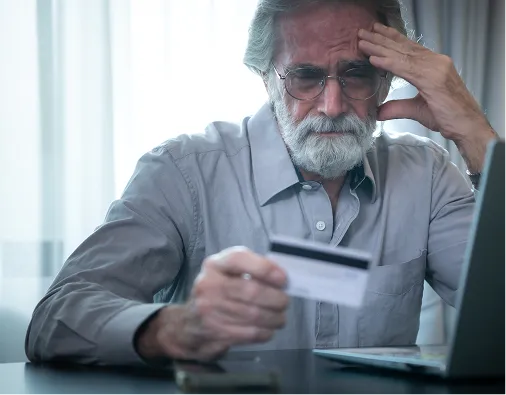 Elderly man with gray hair and beard, wearing glasses, looking worried while holding a credit card near a laptop.