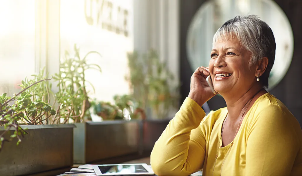 Smiling mature woman in yellow sweater talking on phone near window with plants.