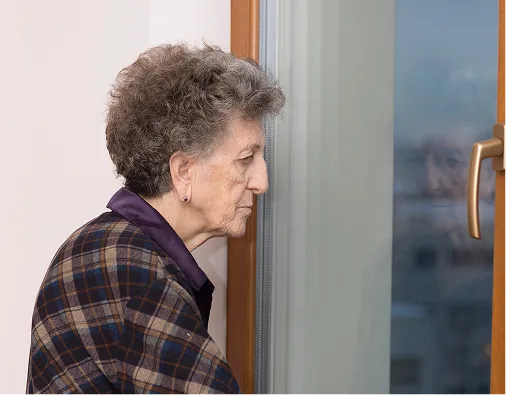 Elderly woman with curly gray hair wearing a plaid jacket, looking out a window.