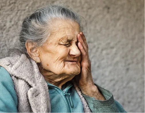 Elderly woman with gray hair and turquoise sweater, touching her face with eyes closed.