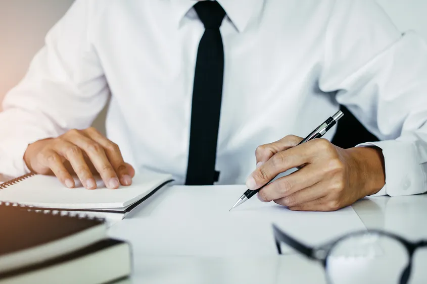 Person in white shirt and black tie writing with pen on blank paper at desk.