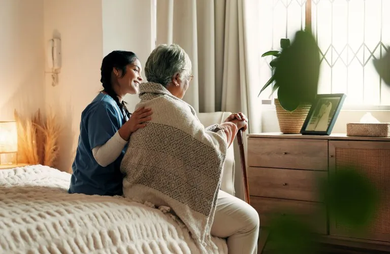 Elderly woman with cane and caregiver sitting on bed, looking at photo frame near window.