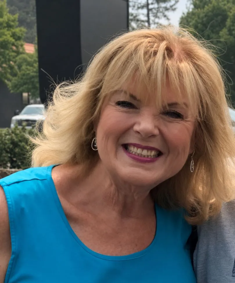 Smiling middle-aged woman with blonde hair wearing a blue sleeveless top and silver earrings outdoors.