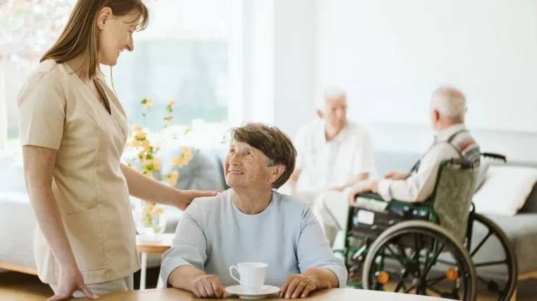 Caregiver interacting with elderly woman at table, two elderly men in background.