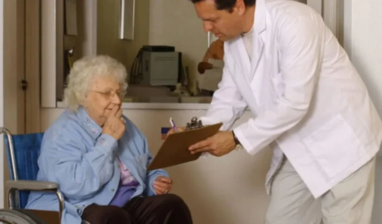 Elderly woman in wheelchair consulting with male doctor holding clipboard in medical office.