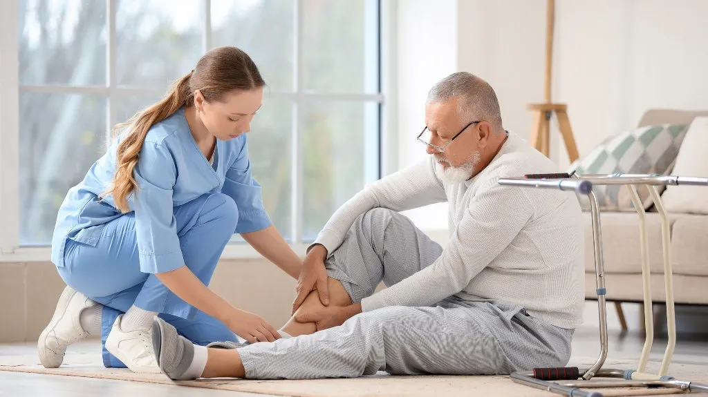 Healthcare professional in blue scrubs examining elderly man's ankle indoors near walker and sofa.