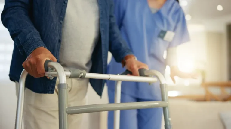 Elderly person using a walker assisted by a healthcare professional in blue scrubs.