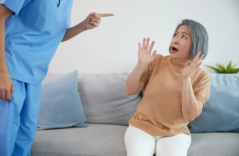 Older woman in beige sweater reacting fearfully to a standing person in blue medical scrubs.