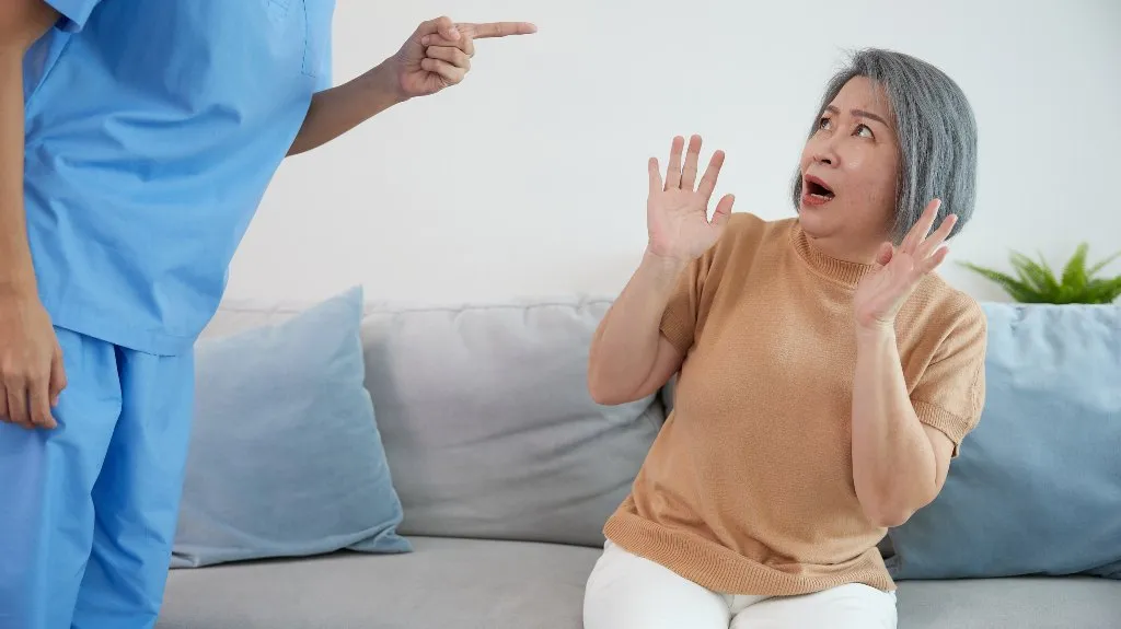 Healthcare professional in blue scrubs pointing, elderly woman in beige sweater reacting fearfully.