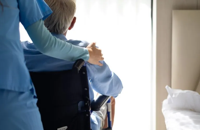 Healthcare professional in blue scrubs supporting elderly patient in wheelchair near bright window.