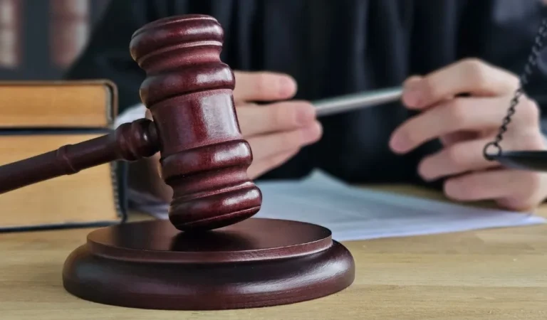 Close-up of a wooden judge’s gavel on a desk with blurred hands holding a pen in the background.