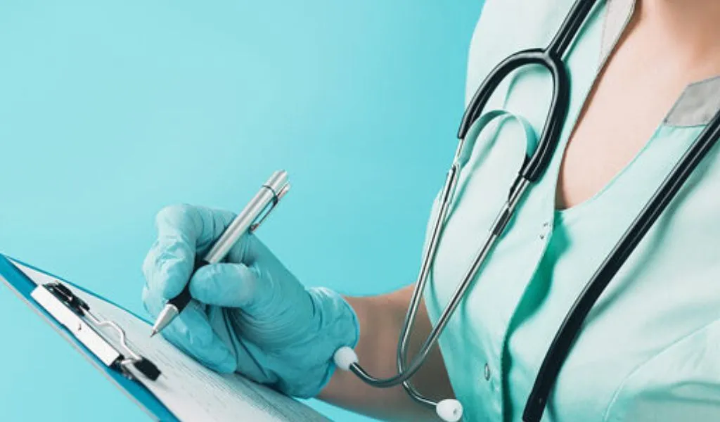 Medical professional wearing teal scrubs and gloves, writing on a clipboard with a stethoscope.