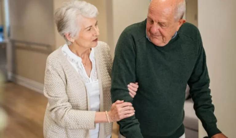Elderly woman supporting elderly man by holding his arm indoors in a home setting.