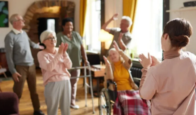 Group of elderly individuals and a caregiver engaging in seated and standing exercises indoors.