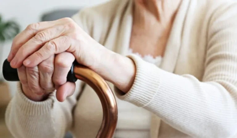 Close-up of elderly person’s hands gripping a brown cane, wearing a beige sweater.