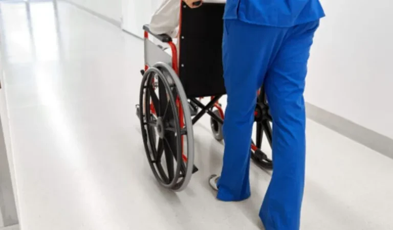 Healthcare worker in blue scrubs pushing an empty wheelchair in a hospital corridor.