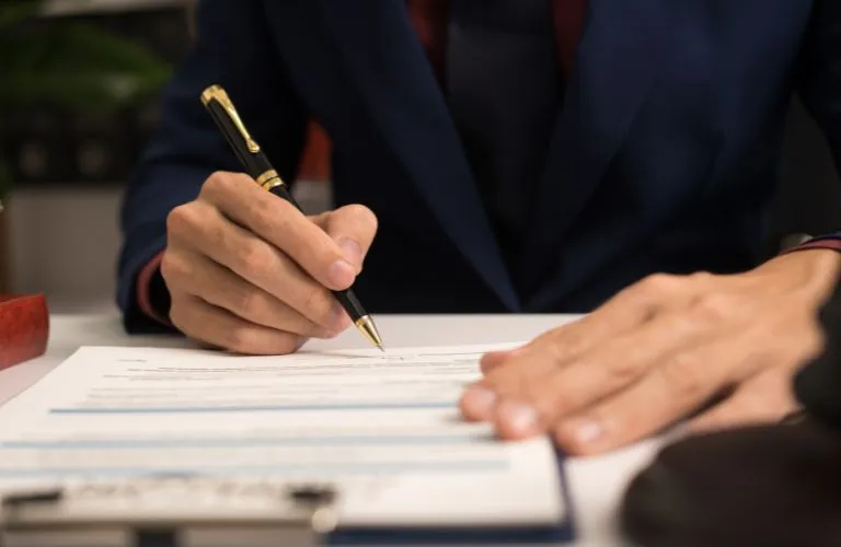 Person in dark suit signing a document with a black and gold pen on white paper.