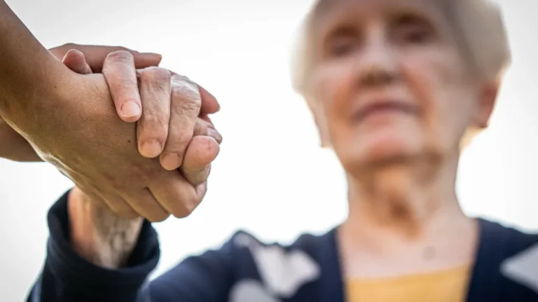 Close-up of two hands clasped, with an elderly person blurred in the background.