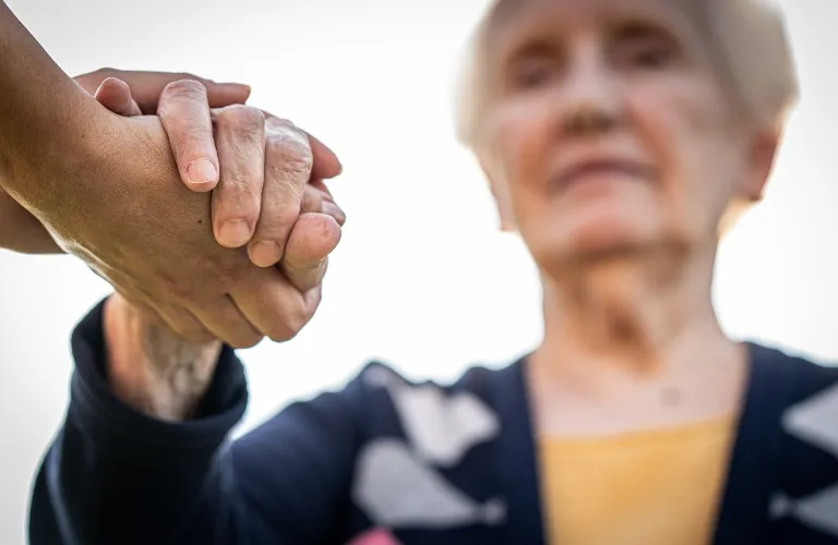 Close-up of a younger hand gently holding an elderly person's hand, blurred elderly woman in background.