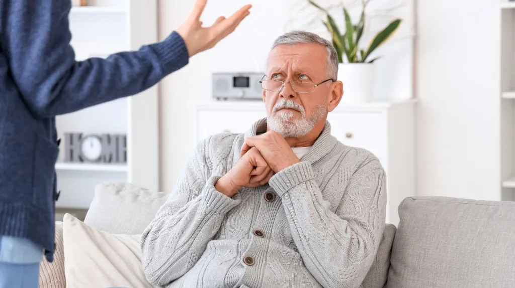 Older man with gray hair and glasses sitting on a gray couch, wearing a beige sweater, looking concerned at a person gesturing with their hand.