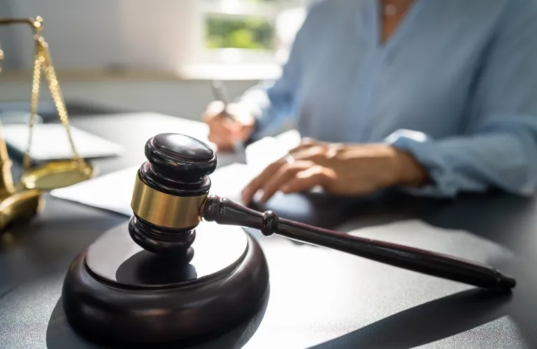 Wooden judge's gavel with brass band on desk, blurred person writing in background.