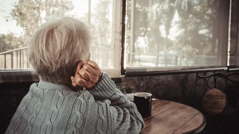 Elderly person with gray hair wearing a gray knitted sweater sitting at a wooden table by a window.