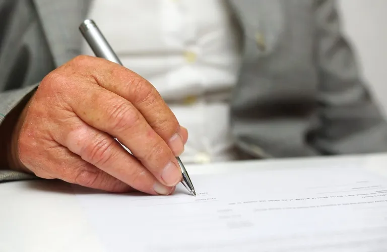Close-up of a person in a gray suit signing a document with a silver pen.