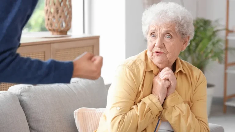 Elderly woman in yellow shirt looking fearful while a person with a clenched fist is visible nearby.