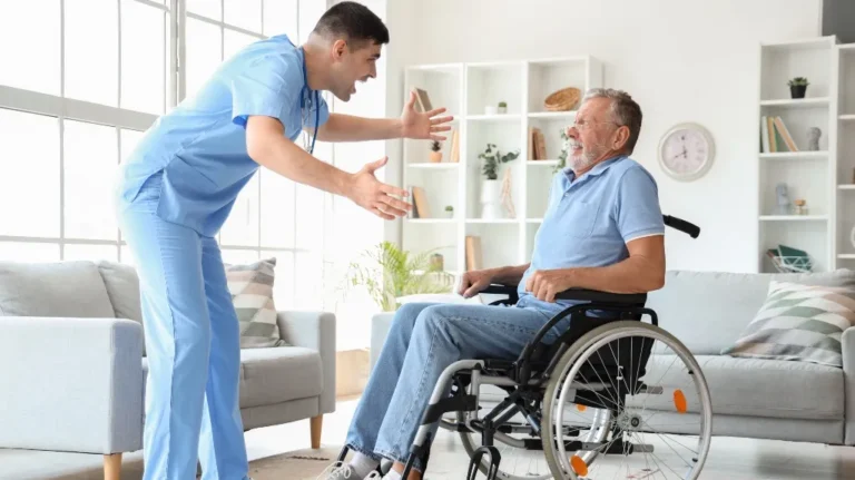Healthcare professional in blue scrubs engaging with elderly man in wheelchair at home.