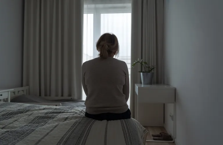 Person sitting on bed facing a window in a dimly lit, minimalist bedroom.