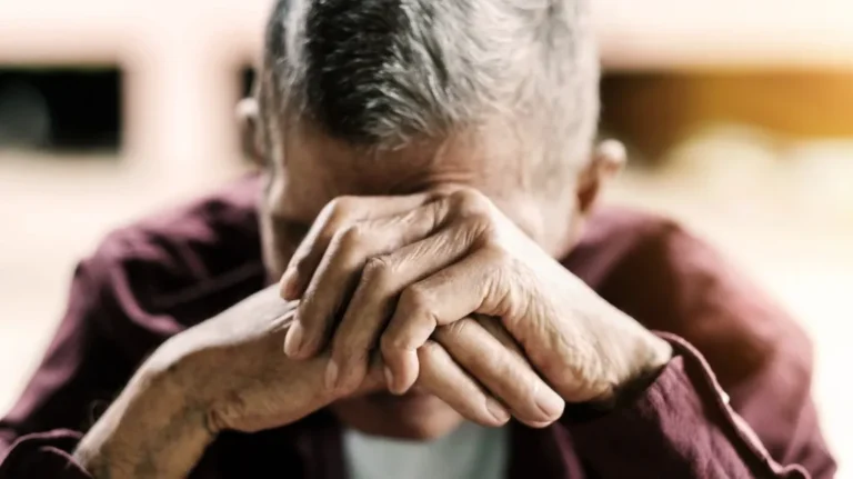 Elderly man with gray hair resting his head on clasped hands, wearing a maroon shirt.
